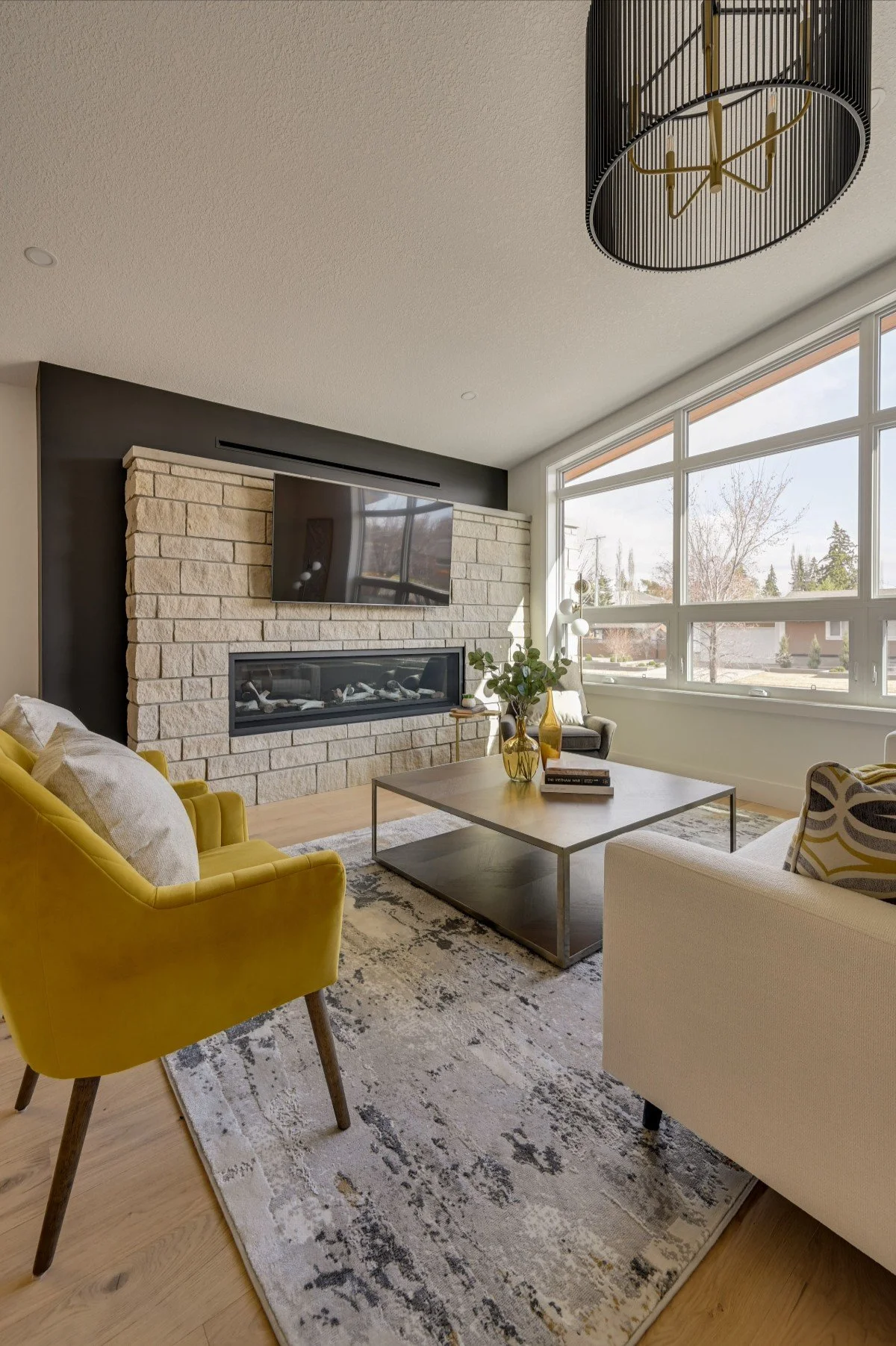Modern living room with a yellow armchair, a white sofa, a rectangular coffee table, a stone fireplace with a mounted TV, large windows letting in natural light, and a black and gold ceiling light fixture.