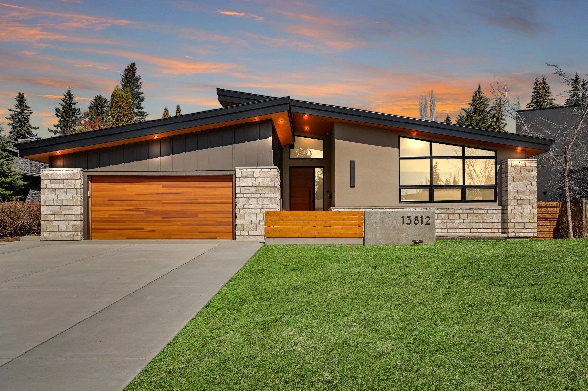 Modern house with a two-car garage, stone and wood exterior, and large front window, sunset sky in the background, and a green lawn in the front yard.