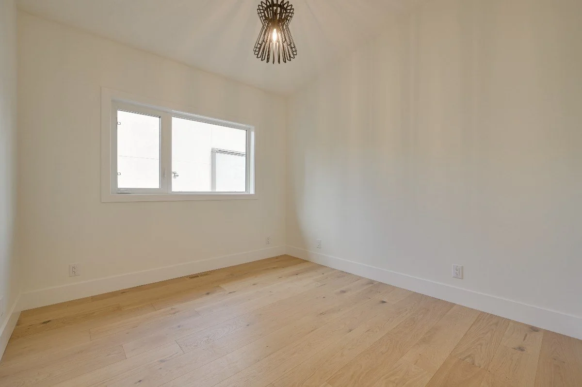 Empty room with wooden floor, white walls, a window, and a modern light fixture hanging from the ceiling.