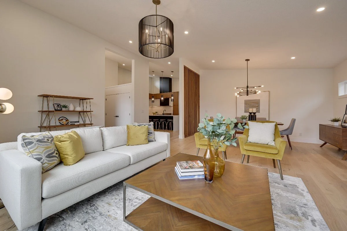 Modern living room with white sofa, yellow armchairs, wooden coffee table with vases and books, wooden flooring, decorative lighting, and artwork on the wall.
