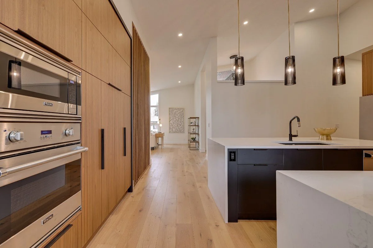 Modern kitchen with wooden cabinets, stainless steel oven, black pendant lights, white countertops, and a sink, opening into a bright living space.