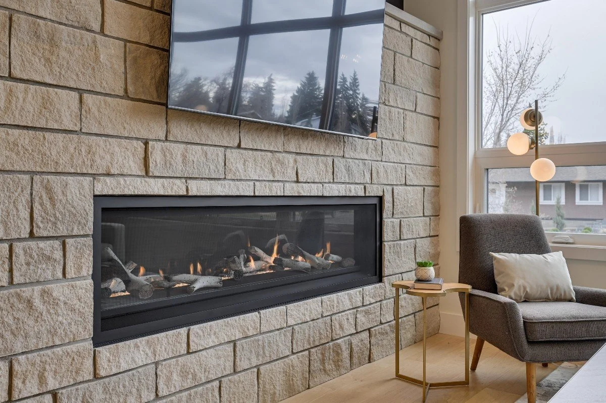 Living room with a stone fireplace, a wall-mounted TV above it, a gray armchair with a pillow, a side table with a small plant and a book, and a floor lamp with three globe lights. There is a large window showing trees and a house outside.