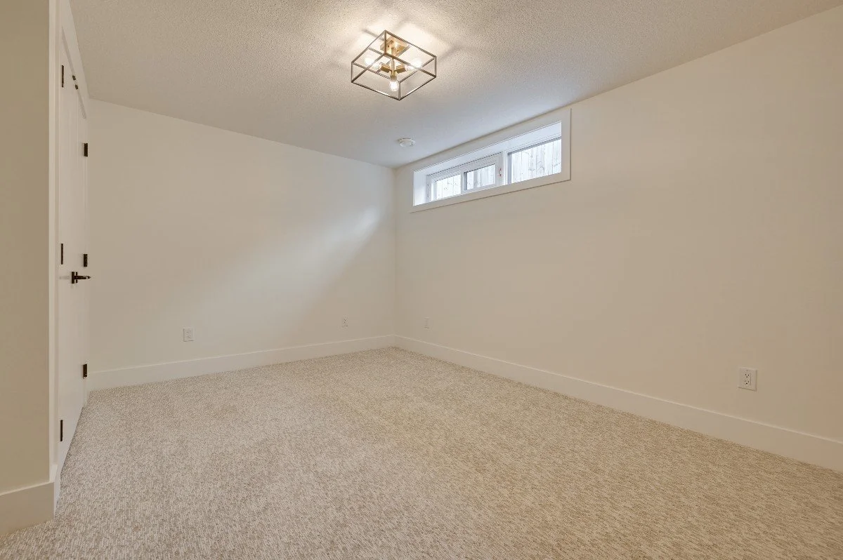 Empty room with white walls, beige carpet, small horizontal window, and modern ceiling light fixture.