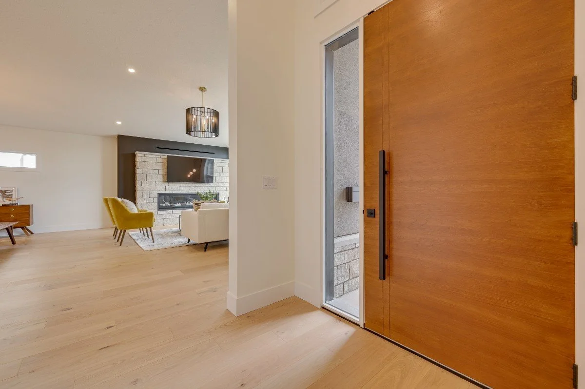 Interior view of a modern home entryway with a large wooden door, a glass storm door, and a glimpse into a living room with light-colored furniture and a fireplace.