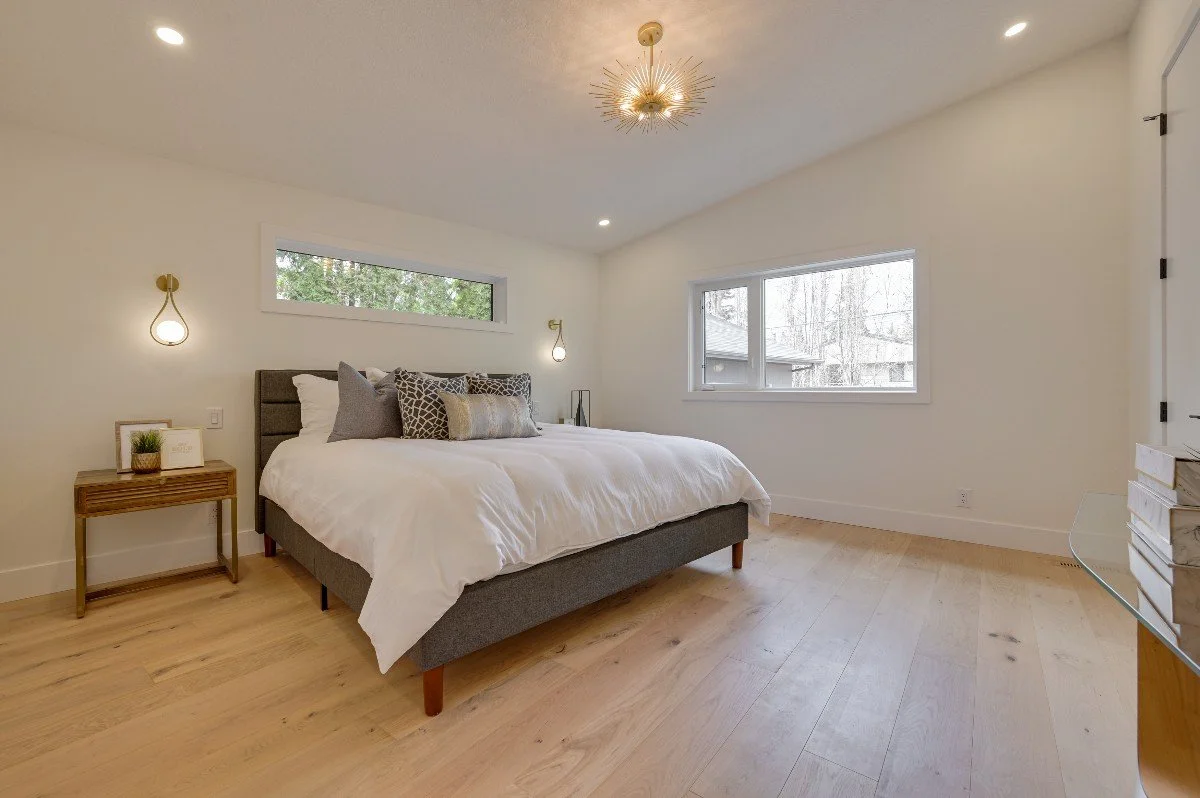 A modern bedroom with white walls, hardwood flooring, a gray upholstered bed with white bedding and decorative pillows, a wooden nightstand with decor items, and two wall-mounted lamps. There are two windows, one high and narrow, the other larger, al