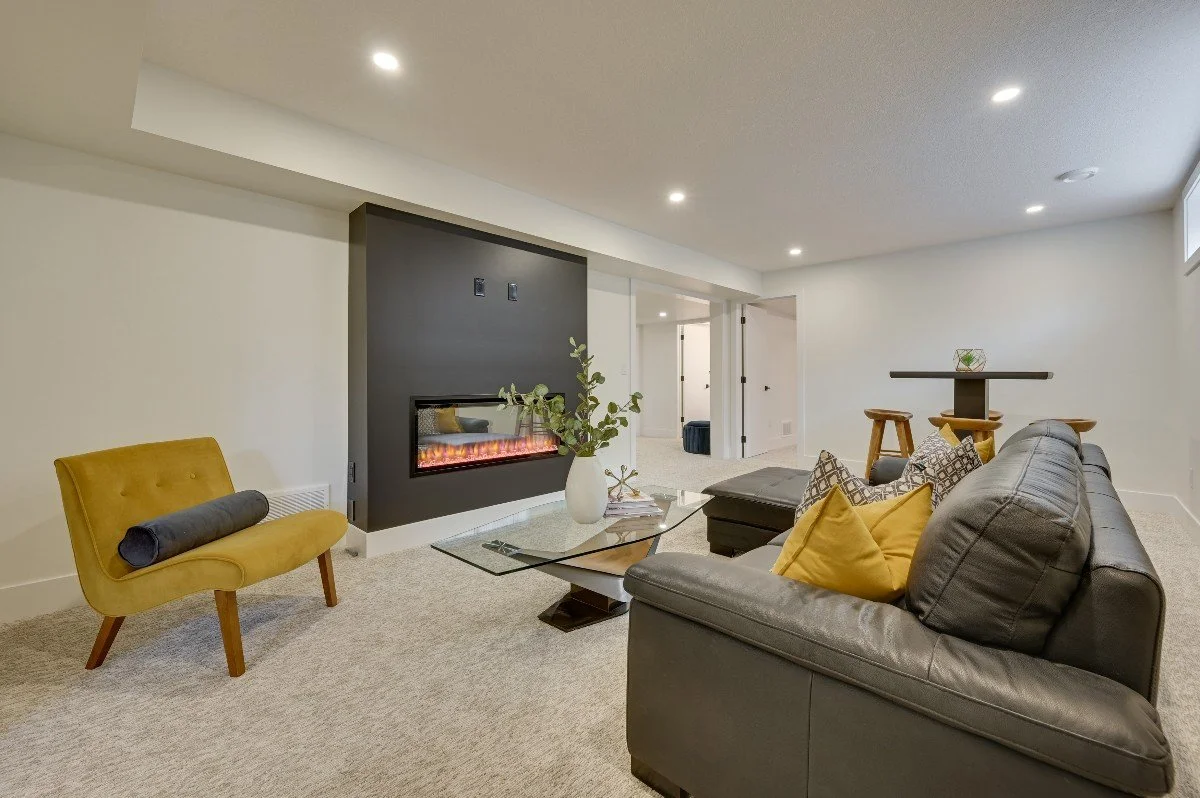 Living room with a gray leather sofa with yellow and patterned pillows, a yellow accent chair with a cylindrical black pillow, a glass coffee table, a modern black fireplace, and a small bar table with stools, all on beige carpeted flooring.