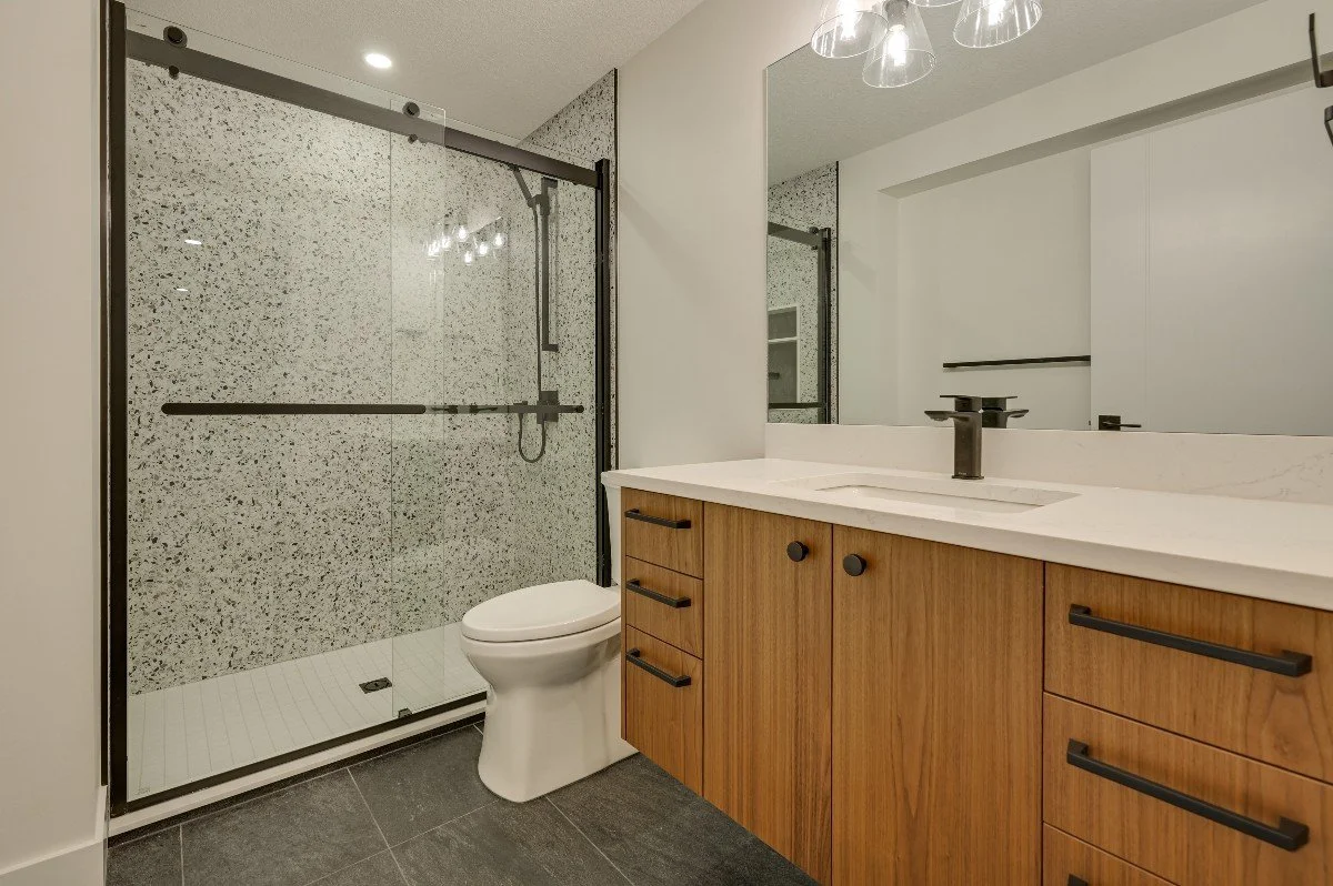 Modern bathroom with a walk-in shower with glass sliding doors, a white toilet, and a wooden vanity with a white countertop and a black faucet. There is a large mirror above the vanity and modern lighting fixtures.