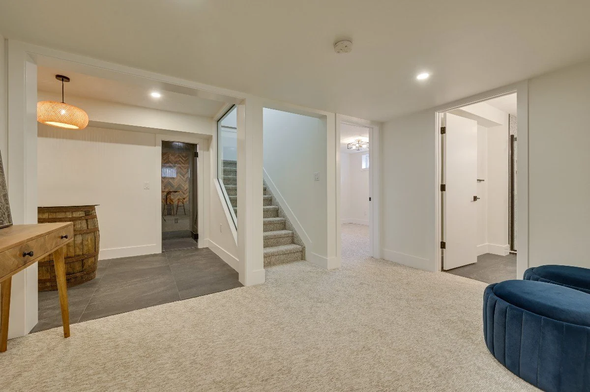 Empty living area with beige carpet, white walls, and doors leading to different rooms, including a staircase with carpeted steps and a small window, with a blue rounded chair in the bottom right corner.