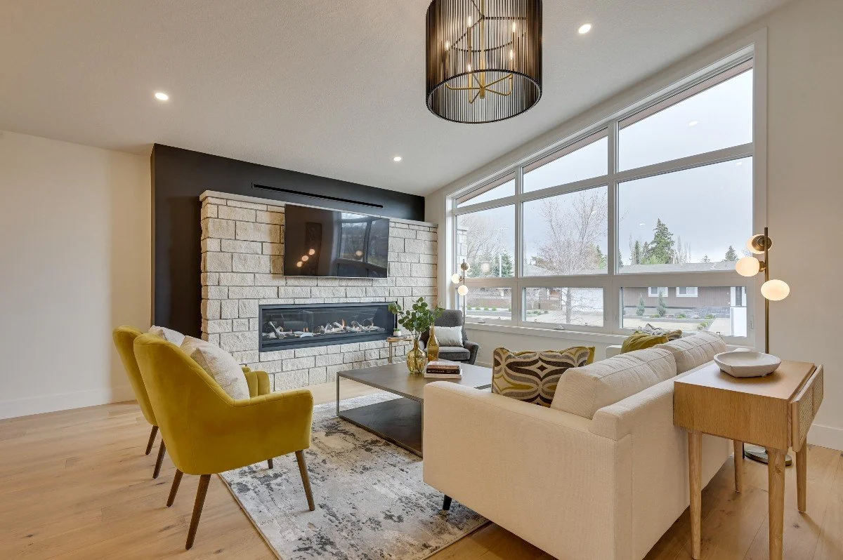 Modern living room with a large window, a stone fireplace, a wall-mounted TV, a beige sofa, yellow accent chairs, a coffee table, and a side table with a lamp.