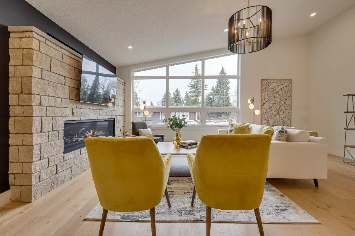 Modern living room with a yellow armchair, a white sofa with pillows, a glass vase with greenery, a large window showing trees outside, and a fireplace with a mounted flat-screen TV above it.