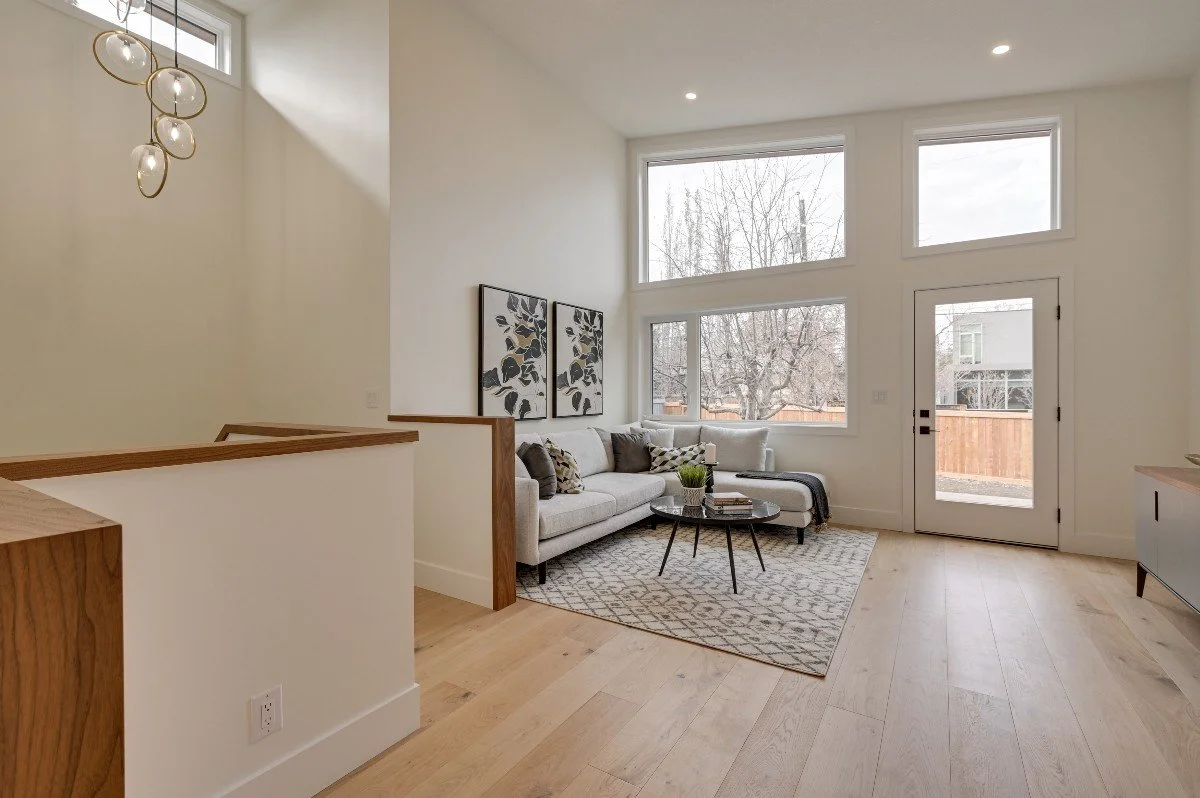 Living room with white walls, large windows, light wood flooring, a white sectional sofa with patterned pillows and a throw blanket, a round black coffee table with books and a plant, a patterned area rug, and wall art with black and gold leaf design