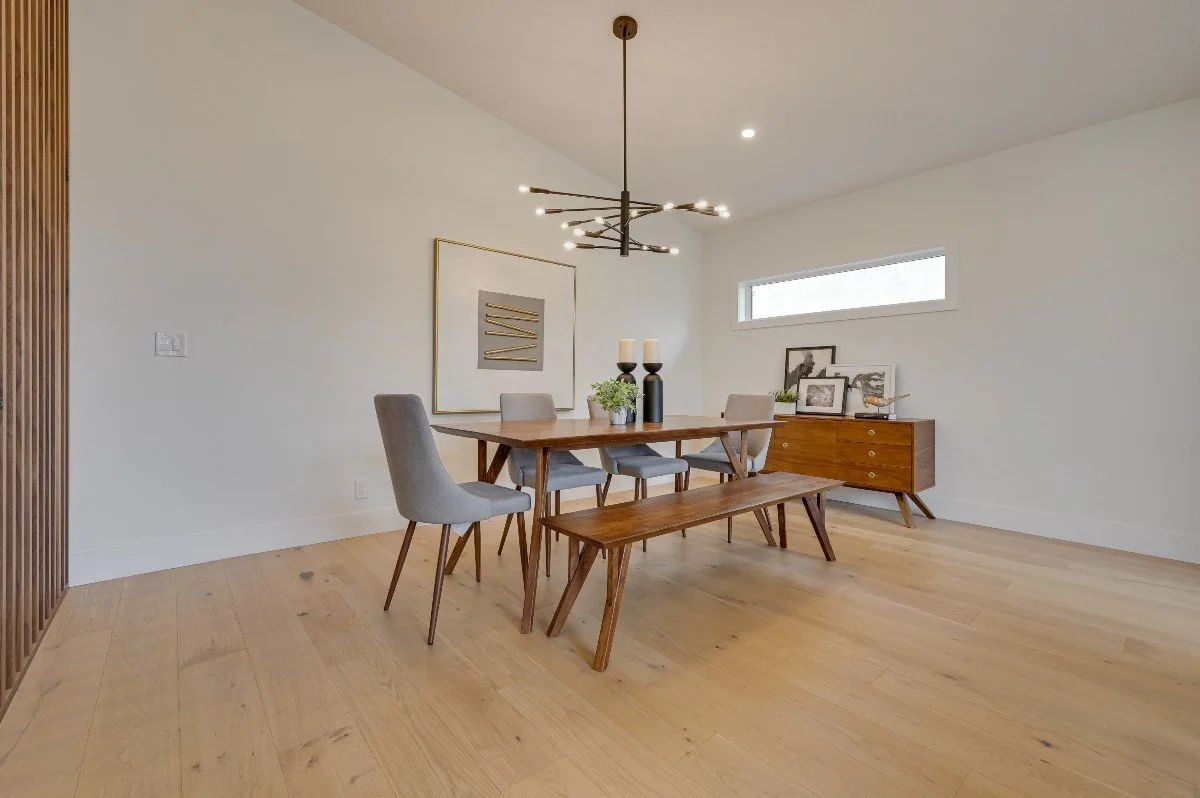 Modern dining room with a wooden table, six chairs, a bench, and a sideboard, decorated with framed pictures, vases, and candlesticks, with a horizontal window and a contemporary chandelier.