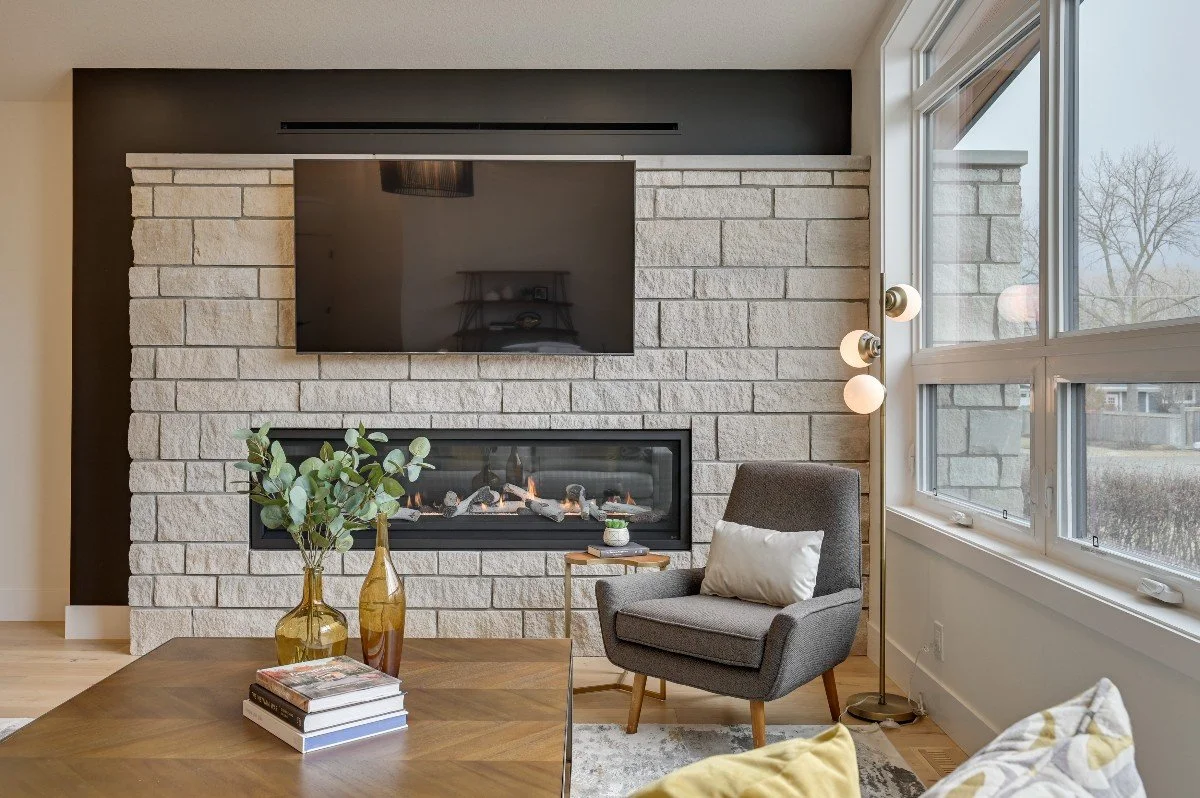 Interior of a living room with a stone fireplace, wall-mounted TV, gray armchair with white pillow, floor lamp with illuminated bulbs, window with a view of trees outside, and a wooden coffee table with vases, books, and a small plant.