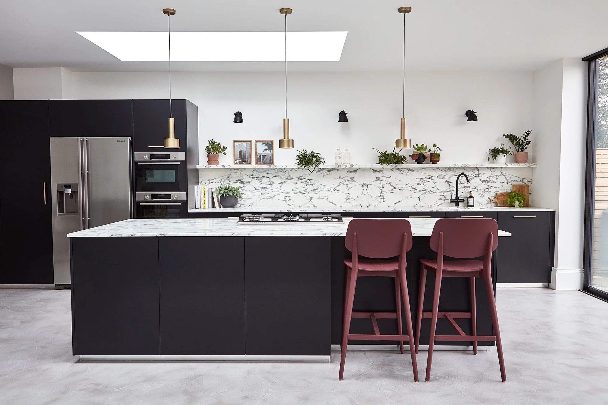 Kitchen design on Trinity Road, Wandsworth. Large dark kitchen with marble worktops and brass pendant lights over the island. Poured concrete effect flooring. Kitchen designed by Stanza interior design, Wandsworth