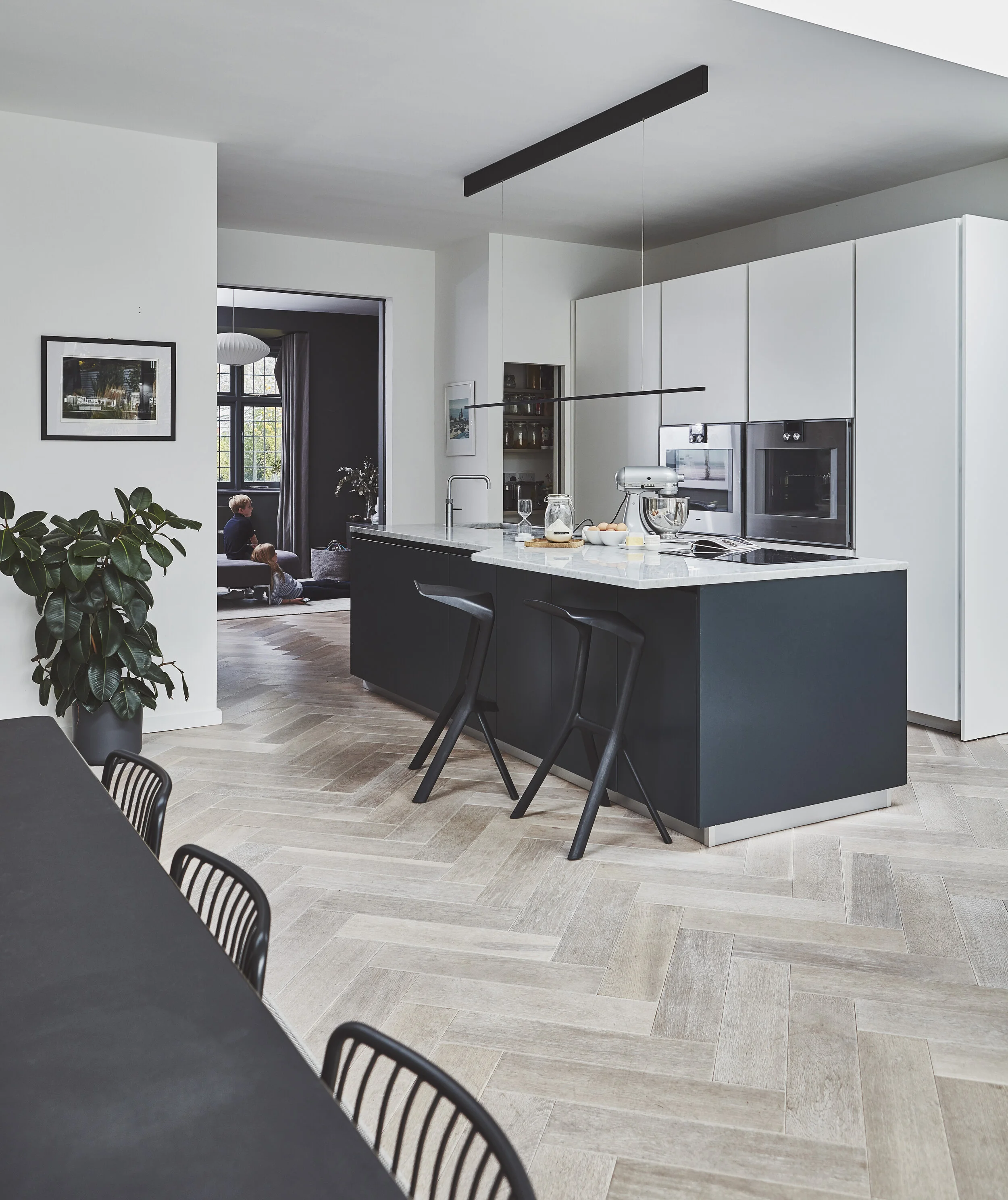 Open plan kitchen and living space with herringbone wood flooring, dark island unit and walk in pantry. Kitchen design by Stanza interior design, Wandsworth Common