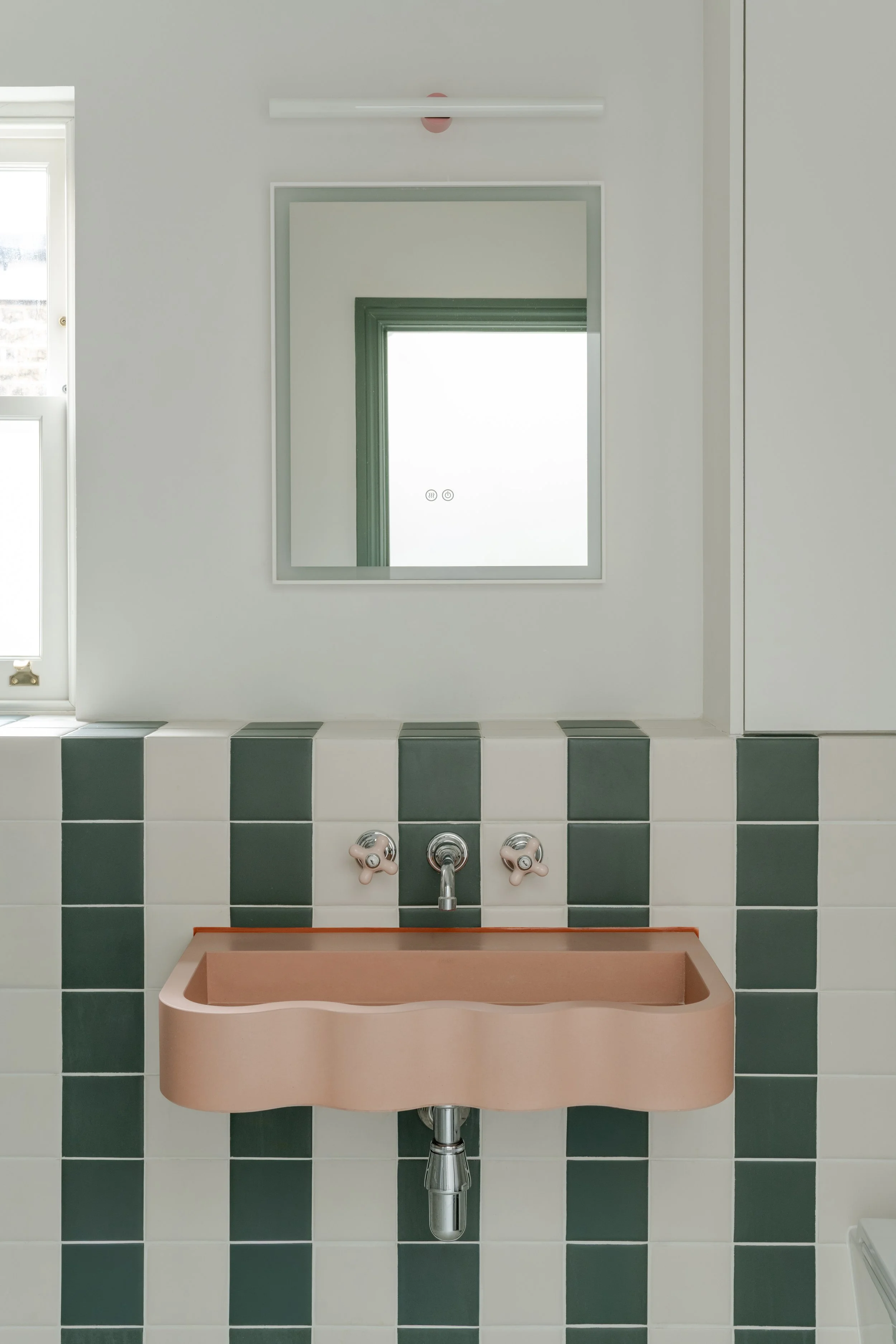 wavy-edged terracotta pink wall-mounted sink, set against vertical stripes of dark green and cream square tiles.