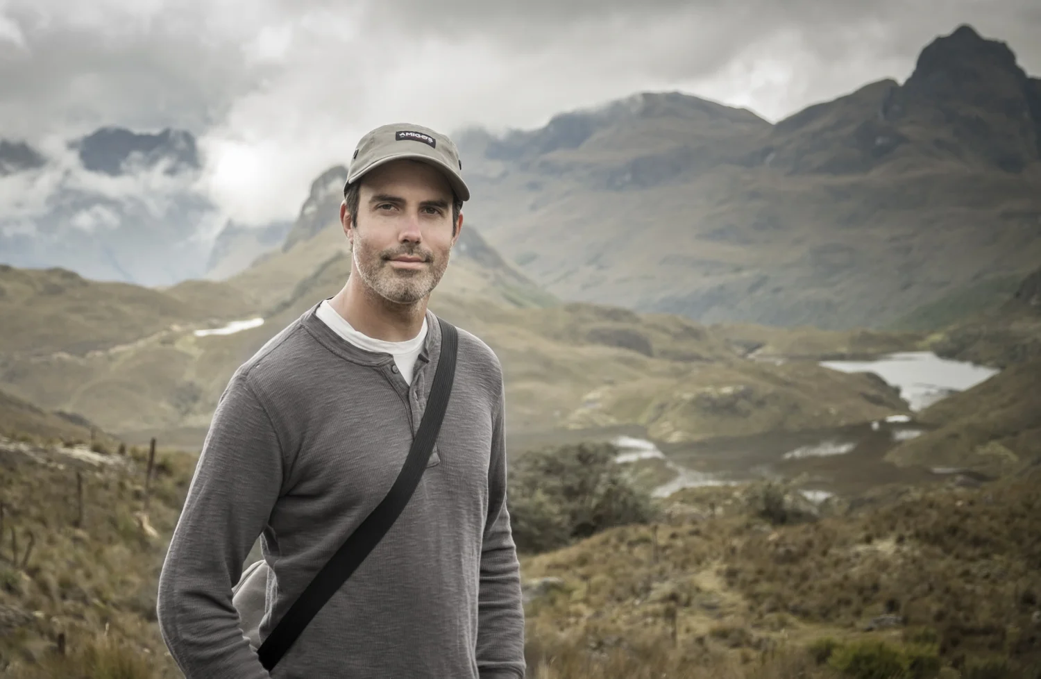 A man standing outdoors in a mountainous landscape with cloudy skies, wearing a gray long-sleeve shirt, a beige cap, and carrying a black strap across his chest.