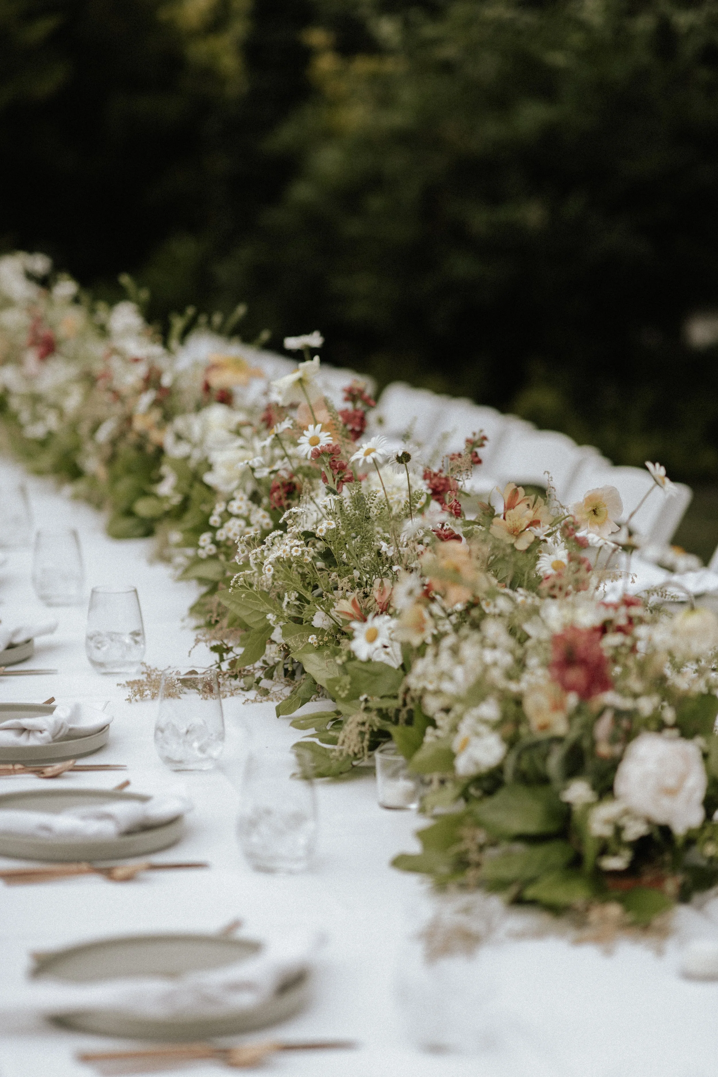 Long table with white tablecloth decorated with a large floral arrangement and set with plates, glasses, and silverware for an outdoor event.