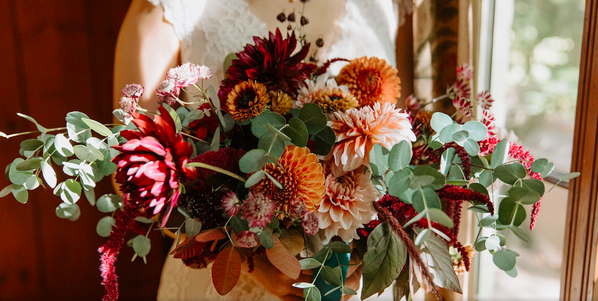 A person holding a large bouquet of colorful flowers, including dahlias, chrysanthemums, and foliage, near a window.