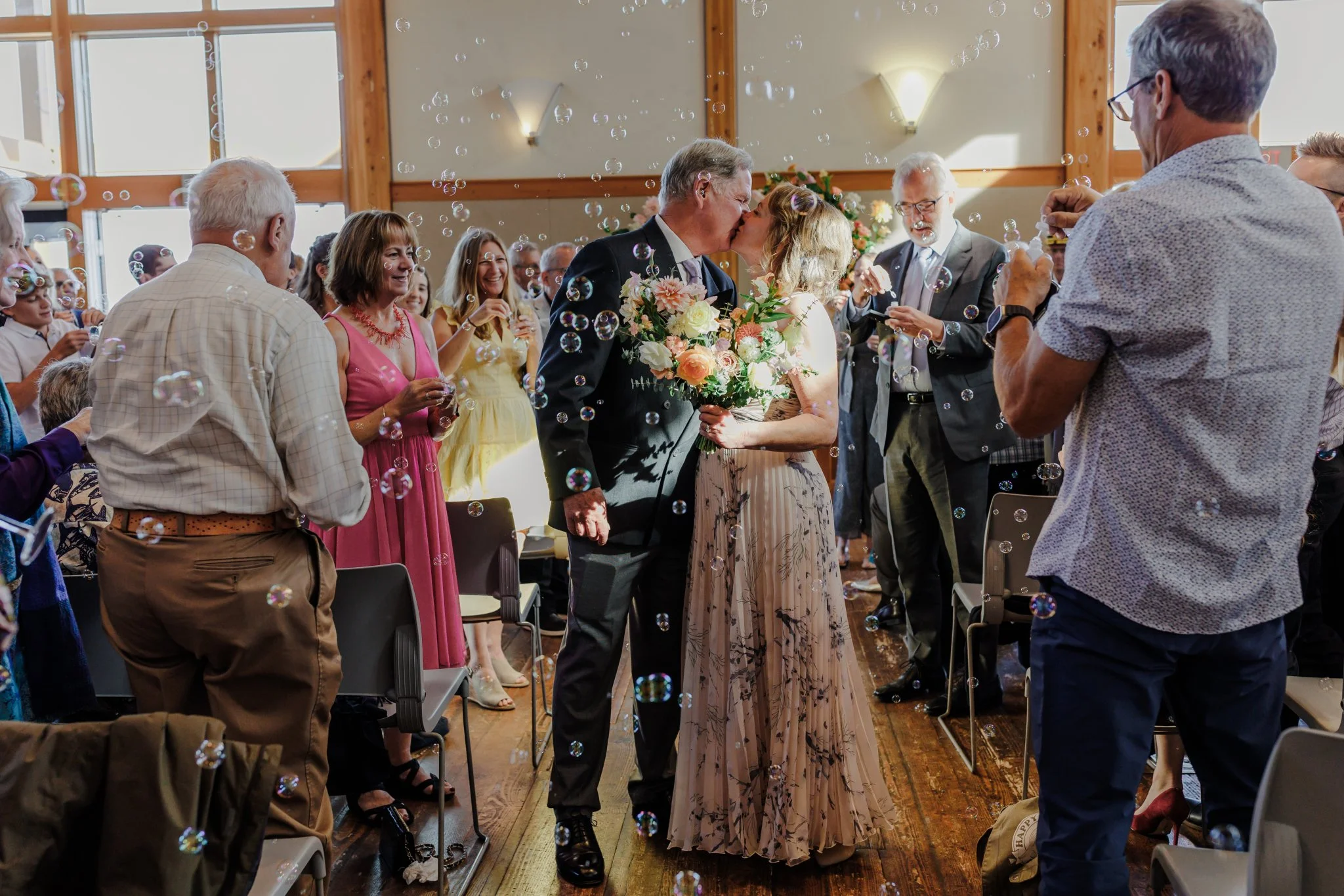 A wedding ceremony inside a decorated venue where a bride and groom are kissing. The bride holds a bouquet of flowers, and guests are celebrating and taking photos, with soap bubbles floating around.
