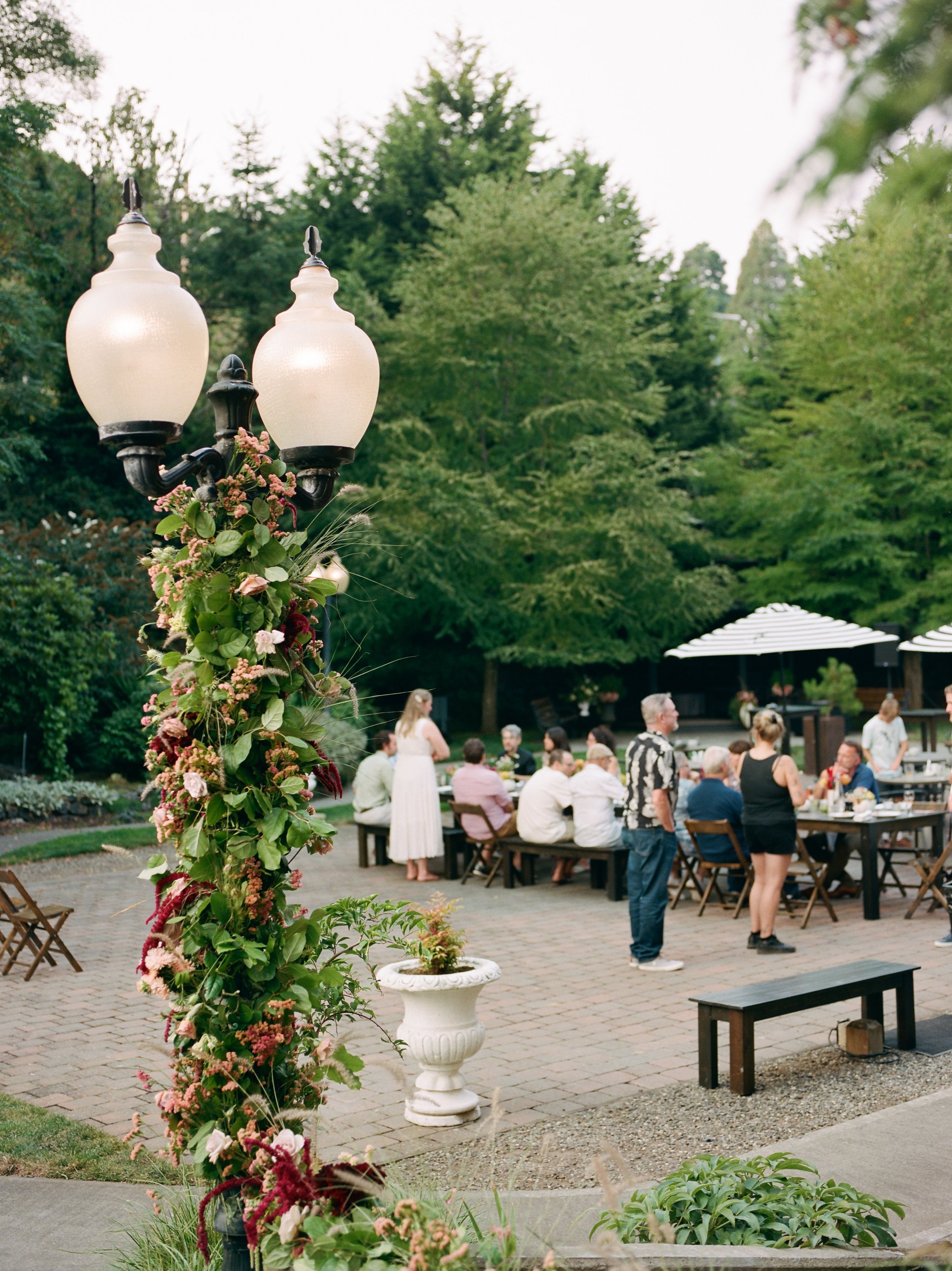 Outdoor gathering with people seated at tables, a person standing, and greenery in the background, along with a decorative streetlamp decorated with flowers.