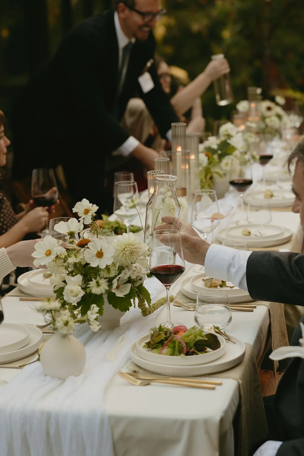 A formal dining table set for a celebration, decorated with white flowers and candles, with guests dressed in formal attire raising glasses of red wine.