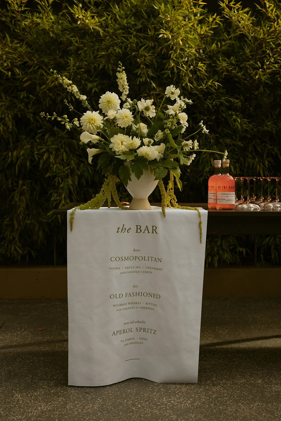 A floral arrangement on a table with a sign listing cocktails, including Cosmopolitan, Old Fashioned, and Aperol Spritz, with bottles of pink drink and glassware in the background.