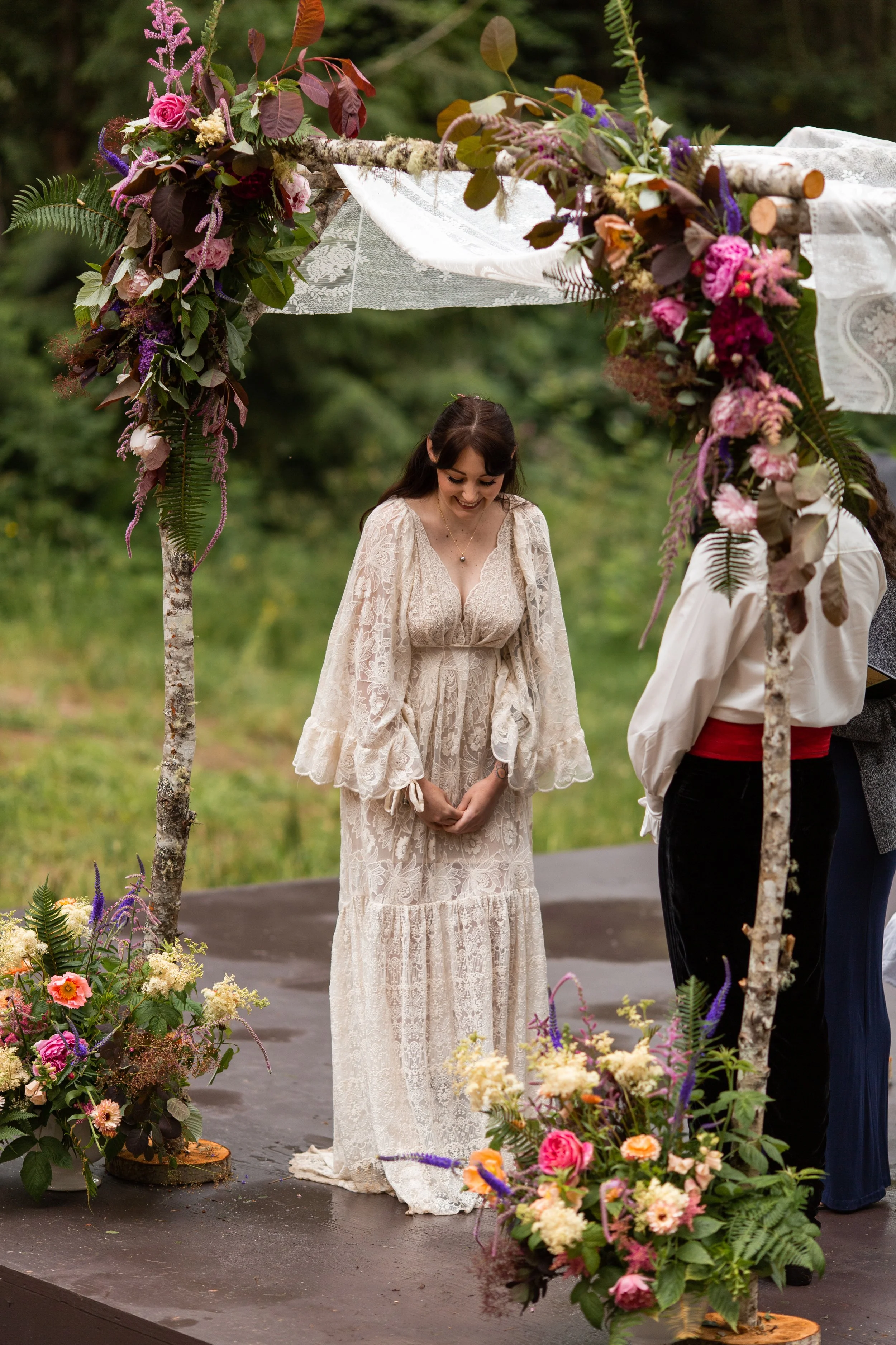 A woman in a long white lace dress standing under a floral arch at an outdoor wedding ceremony, with greenery in the background.