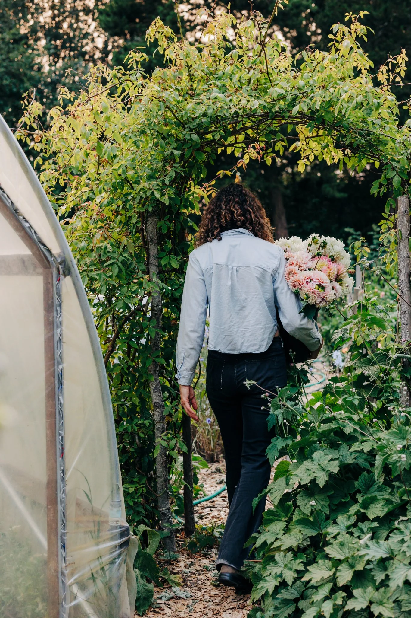 A woman with curly hair, wearing a light jacket and dark pants, walking through a green garden path holding a bouquet of pink and white flowers.