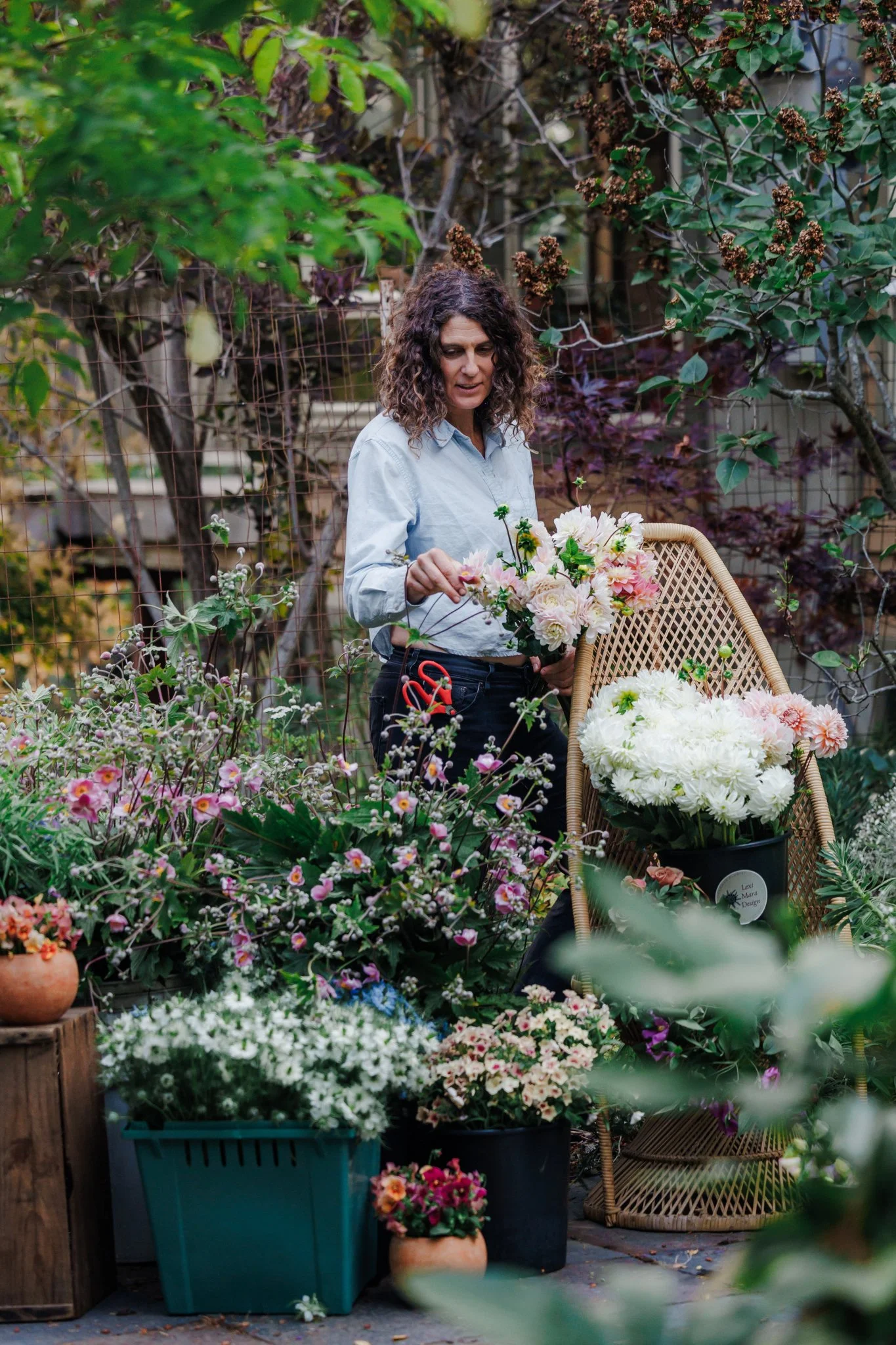 A woman with curly hair in a light blue shirt arranges flowers in a garden with abundant plants and colorful blossoms.