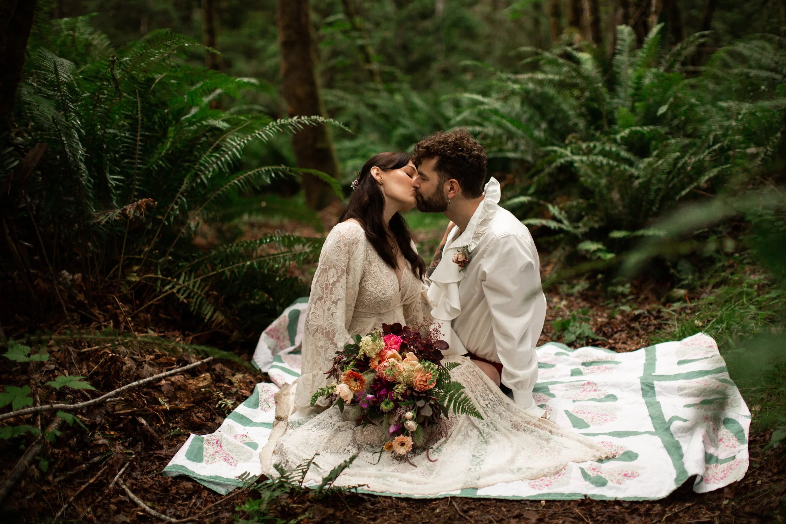 A couple dressed in vintage wedding attire sharing a kiss while seated on a quilt in a lush forest, with a bouquet of mixed flowers in front of them.
