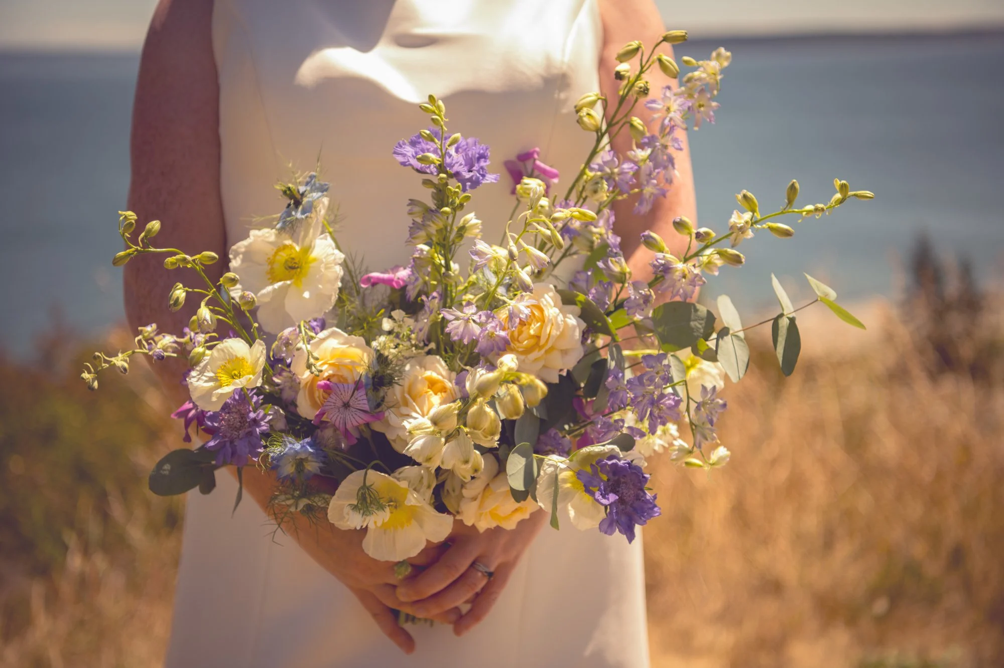 Person holding a bouquet of mixed flowers outdoors, with a field and water in the background.