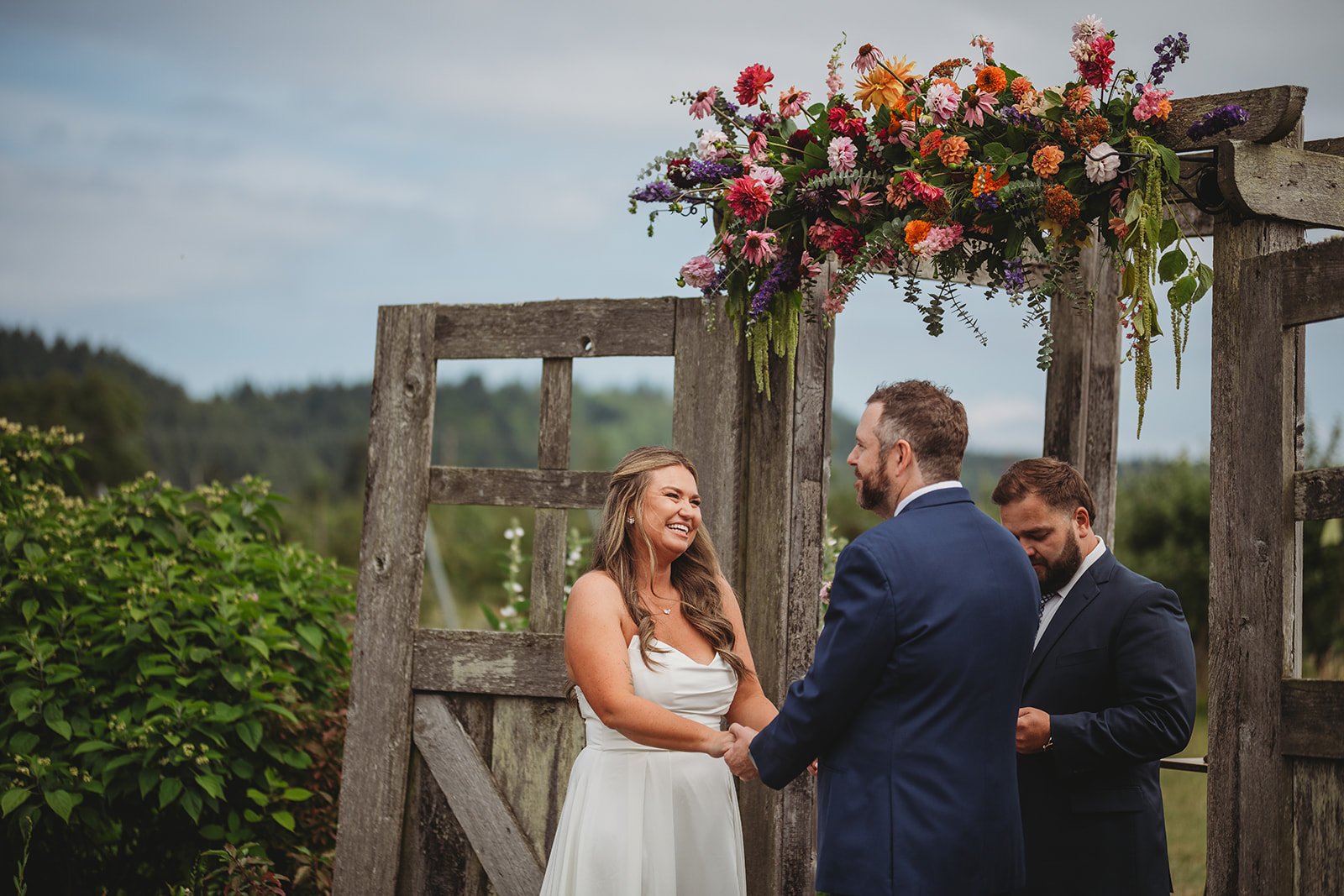 A couple getting married outdoors, holding hands and smiling at each other, with an officiant standing beside them, under a rustic wooden arch decorated with colorful flowers.