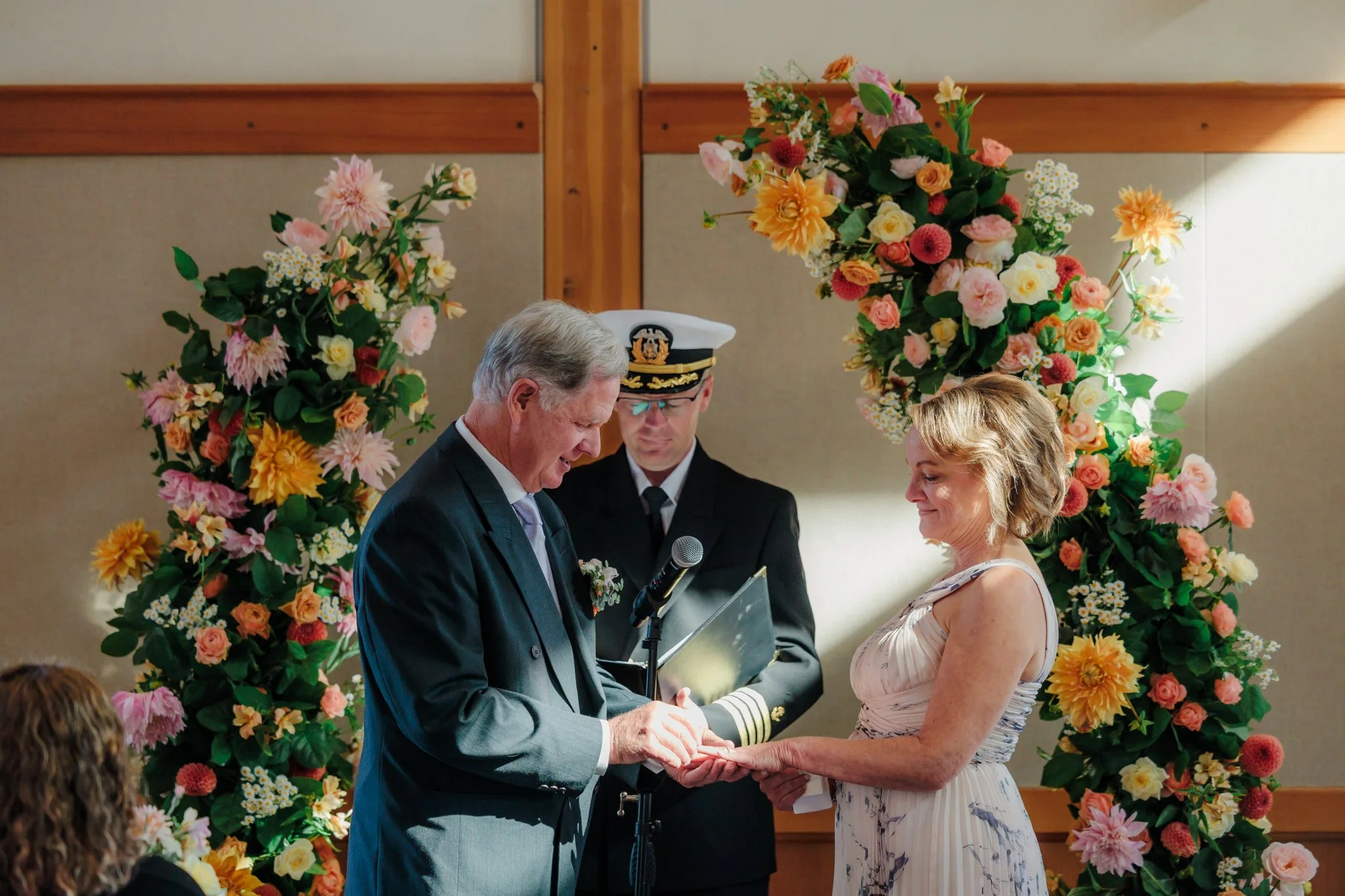 An elderly man in a gray suit and tie receiving an award or recognition from a woman in a sleeveless dress, with a man in a naval officer uniform standing behind them. They are in front of a floral arch with pink, yellow, and orange flowers at a formal ceremony or celebration.
