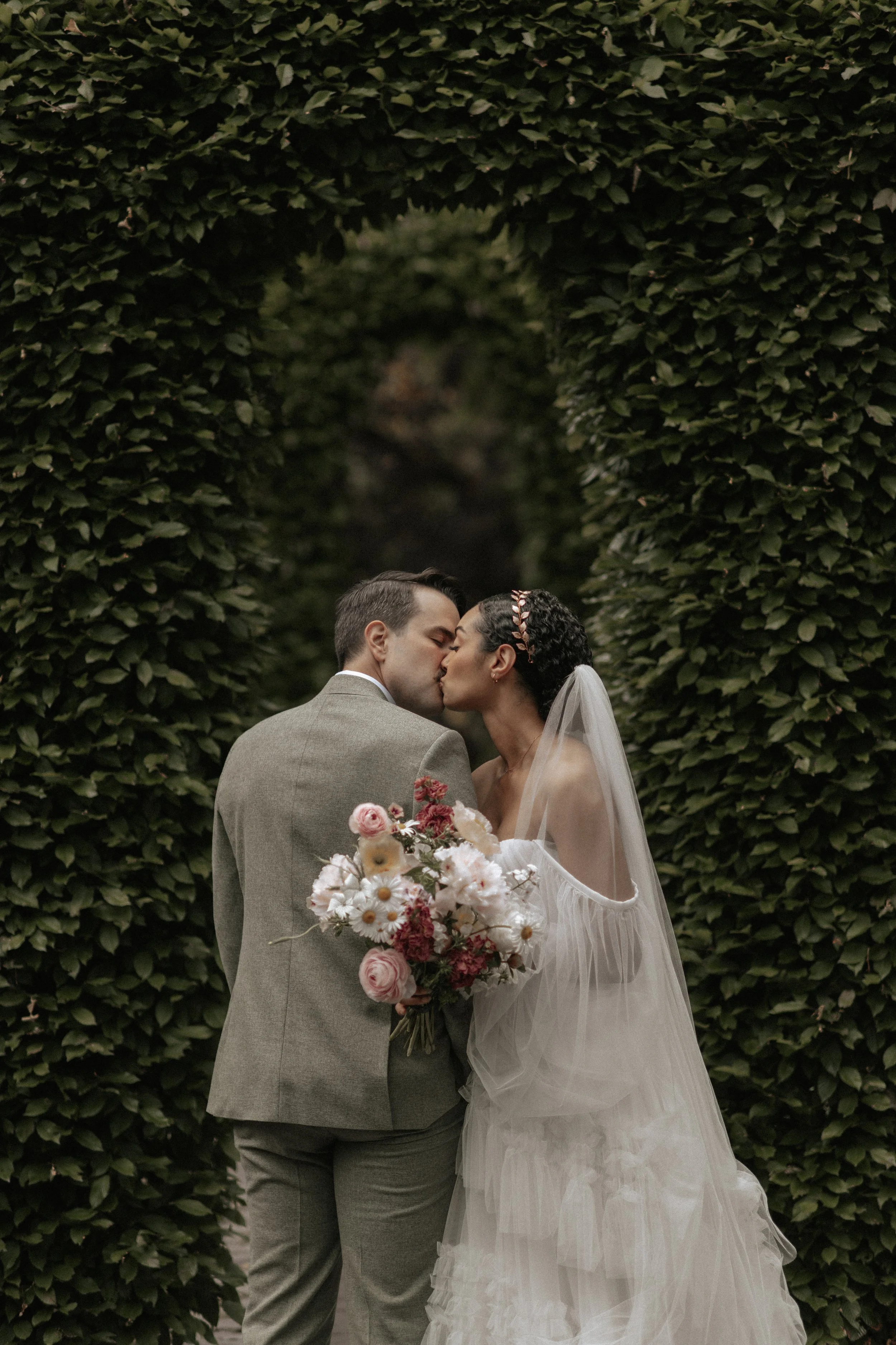A bride and groom kiss in an outdoor wedding, framed by a green leafy arch, with the bride holding a bouquet of pink and white flowers.