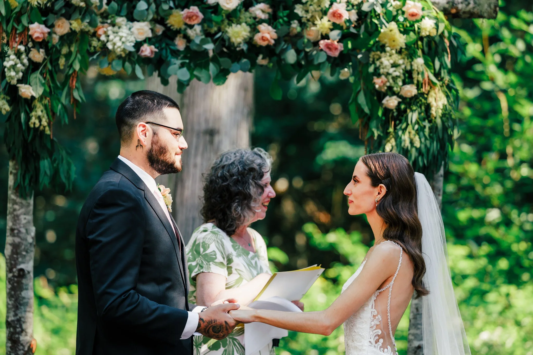 A wedding ceremony outdoors with a bride and groom holding hands, facing each other under a floral arch, with a officiant in the background.