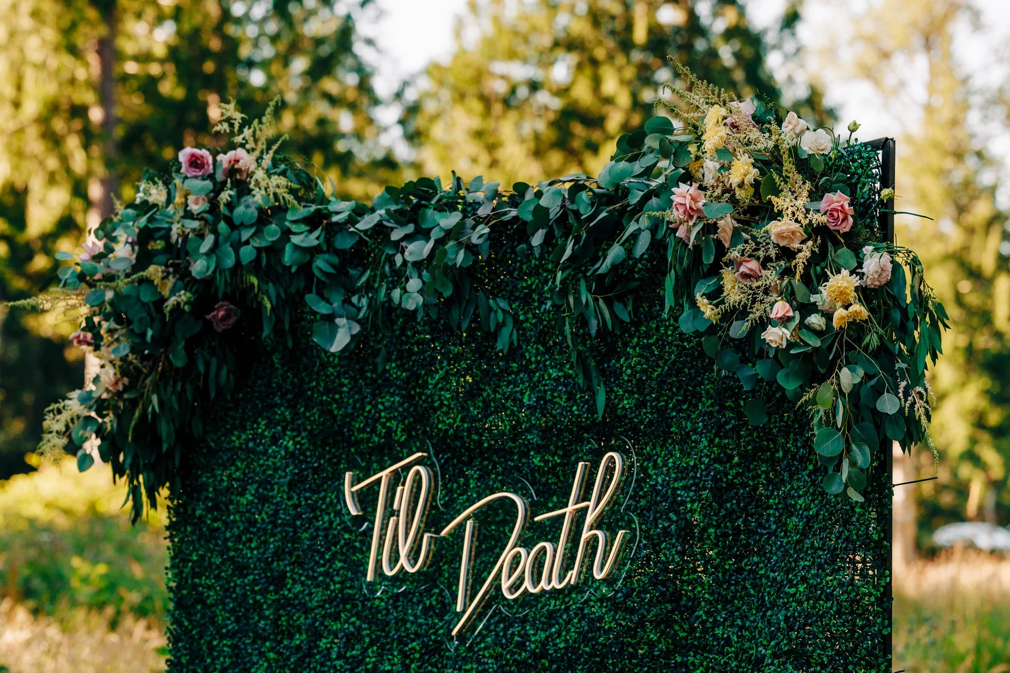 A decorative greenery backdrop with pink and cream flowers and a neon sign that reads 'The Path,' set outdoors with trees and sunlight in the background.