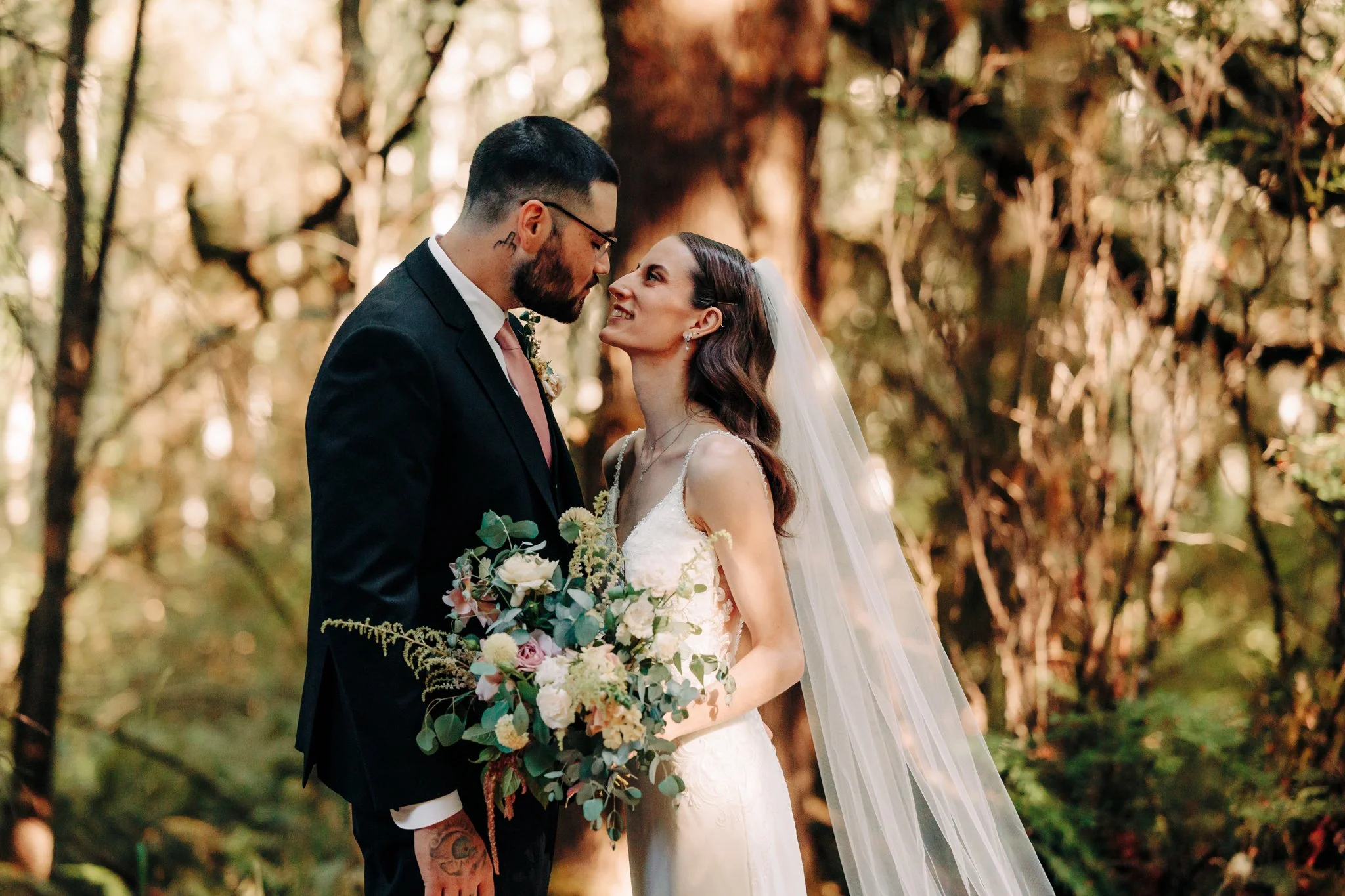Bride and groom standing in a forest, looking into each other's eyes, with the bride holding a bouquet of flowers.
