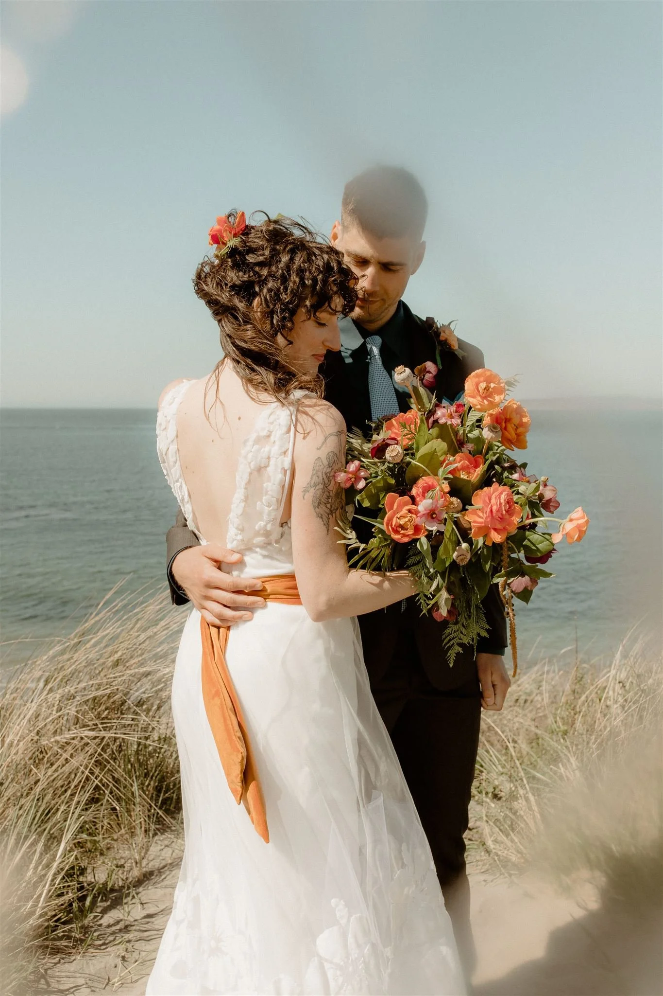 A bride and groom embracing on a beach, with the bride holding a large bouquet of colorful flowers, both dressed in wedding attire