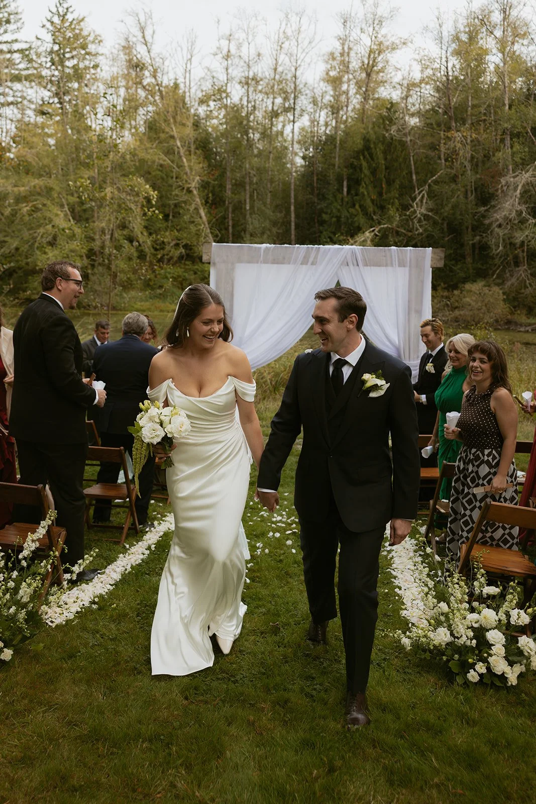 A newly married couple walking down the aisle at an outdoor wedding ceremony, smiling and holding hands, surrounded by guests seated on either side, with trees and a white draped wedding arch in the background.