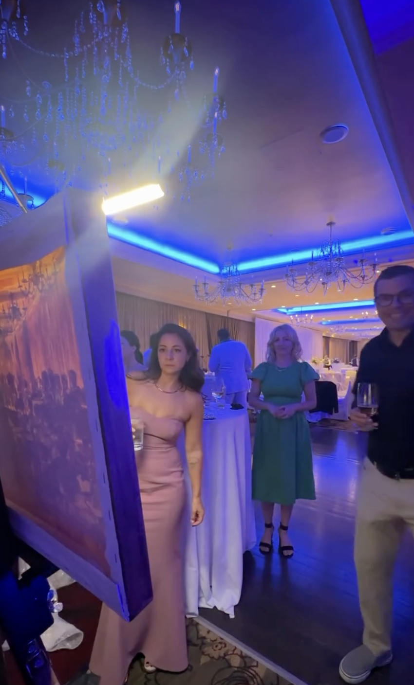  Wedding guest in blush gown pausing beside a large live event painting on easel during a reception in a blue-lit chandelier ballroom 