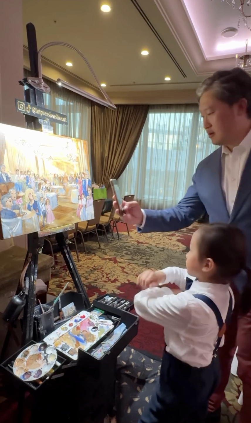  Wedding guests looking at a live reception painting in progress on easel, with artist palette and supplies visible in the foreground 
