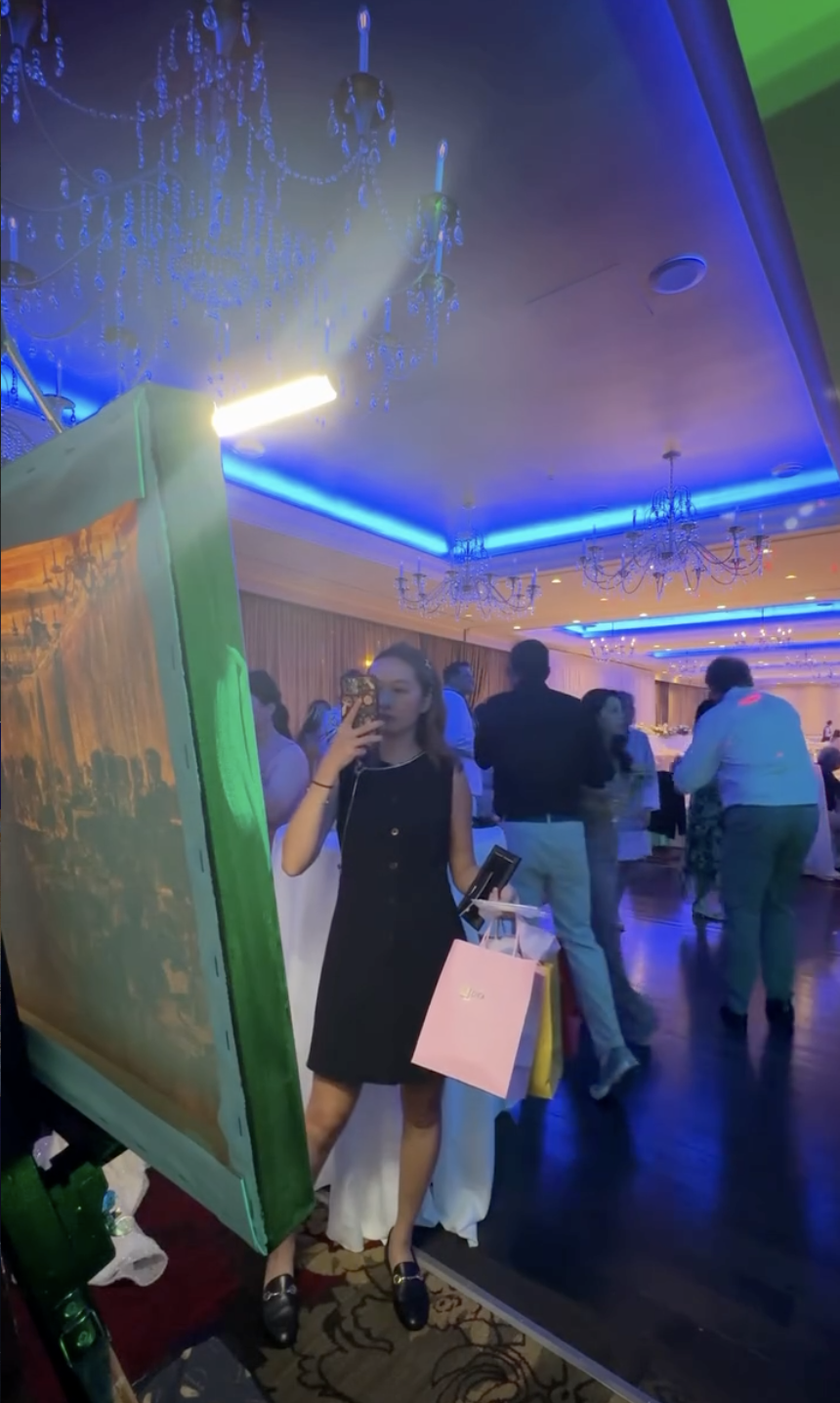  Wedding guest taking a photo of a large live event painting on easel in a chandelier ballroom with blue ambient lighting 