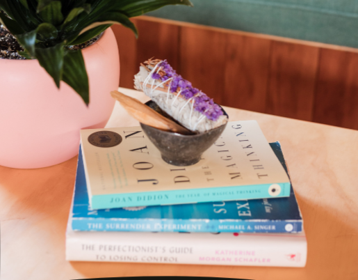 Close-up of a cozy living room corner with a potted plant, stack of books, a smudging stick, and a decorative pillow on a teal sofa.