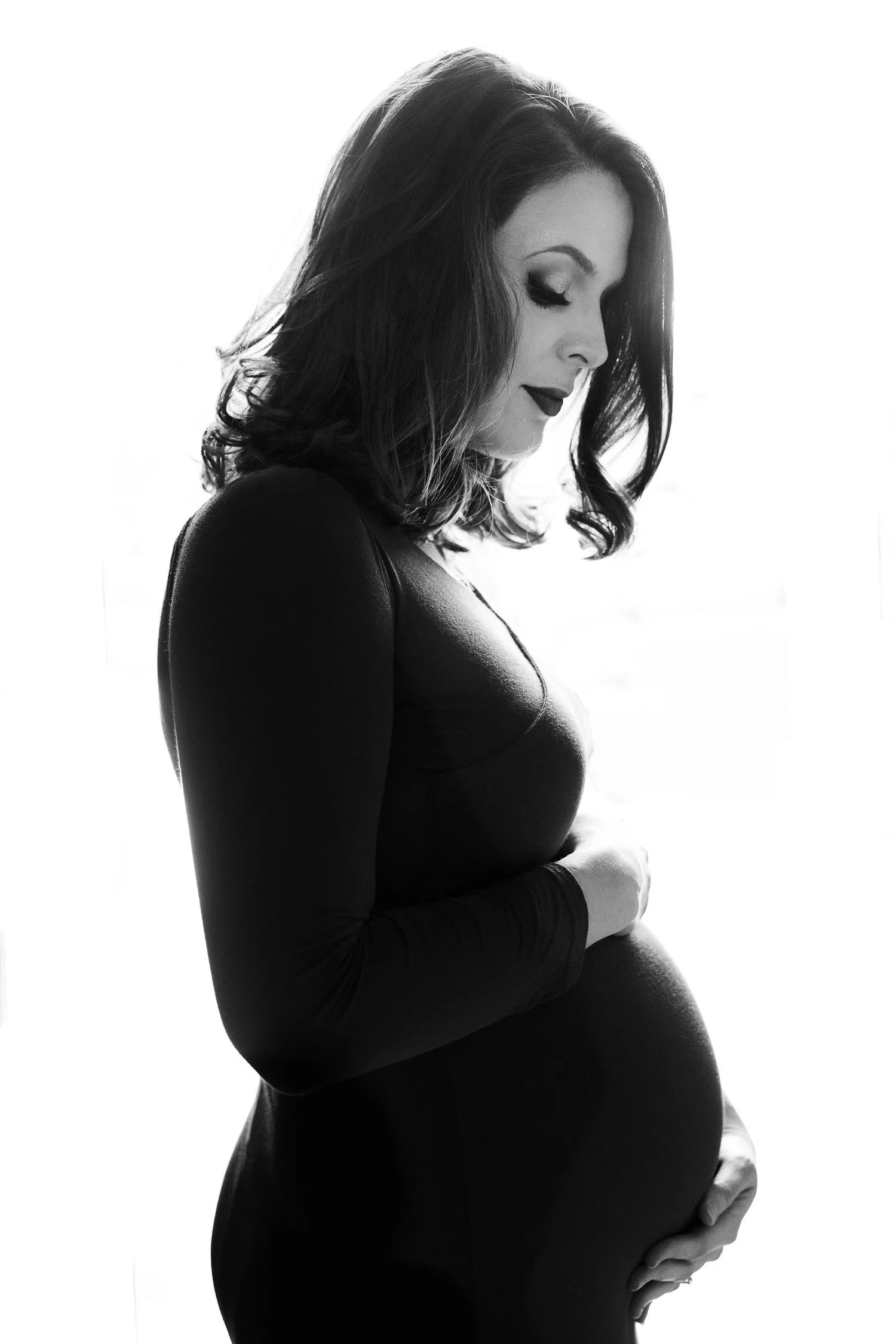 Black and white photo of pregnant woman with shoulder-length hair, looking down, gently holding her belly with both hands in Lees Summit MO.