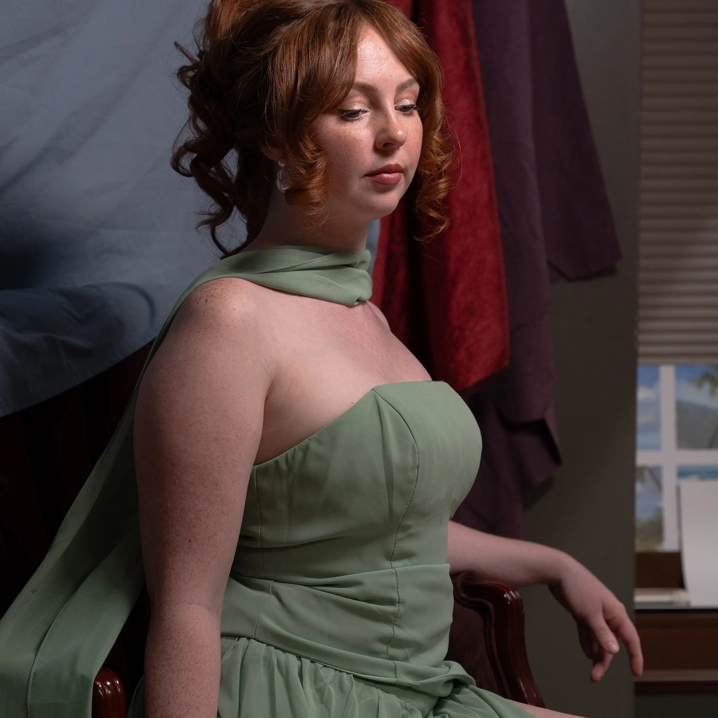 A woman with red hair and freckles wearing a sleeveless green dress, sitting in a room with red and purple curtains, looking downward.