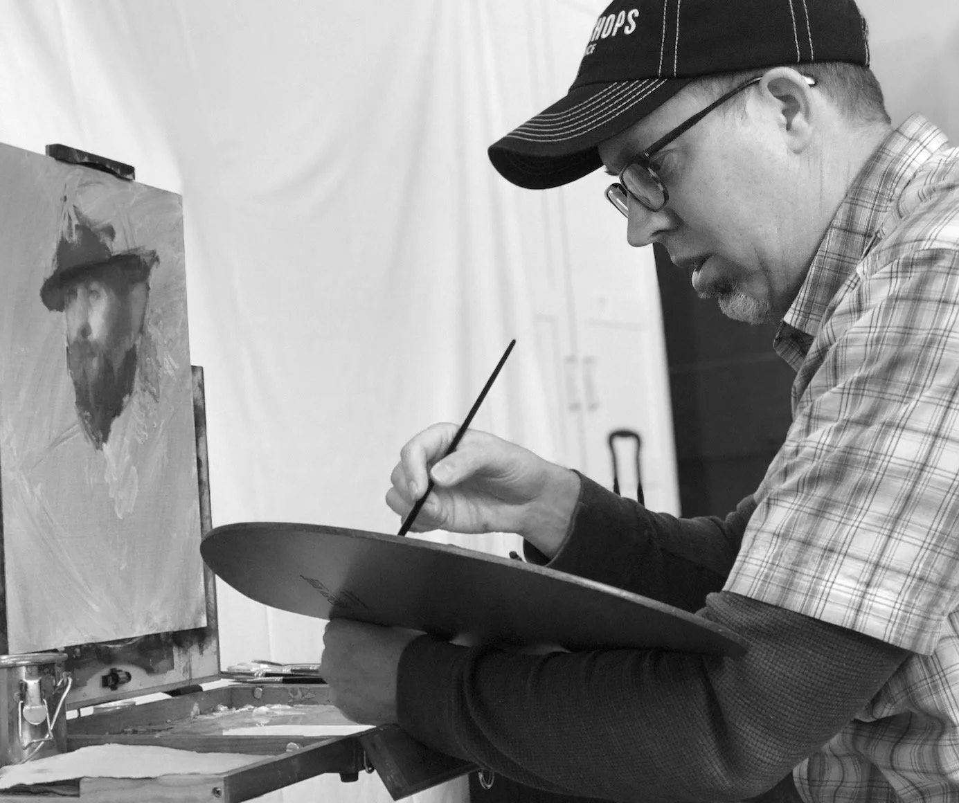 Casey Childs is painting on a canvas, holding a palette and a brush, with an easel and art supplies nearby.