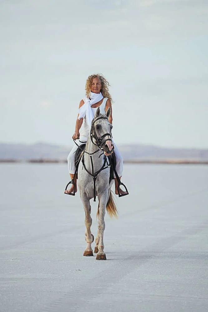 A woman with curly blonde hair riding a white horse across a flat, open landscape with a cloudy sky.
