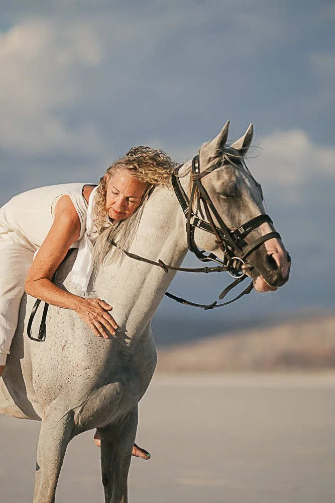 A woman with curly blonde hair hugging a gray horse on a beach with a cloudy sky.