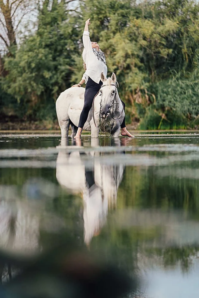 A woman standing on a white horse in a shallow river, with her arm raised upward, surrounded by green trees.
